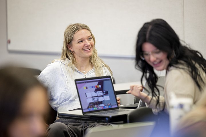 Two female students sit at a table in a classroom smiling as a laptop sits on the table.