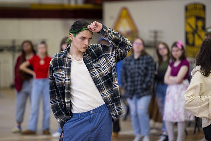 A student-model with black hair and a green streak poses while wearing a flannel shirt, white undershirt and blue pants.