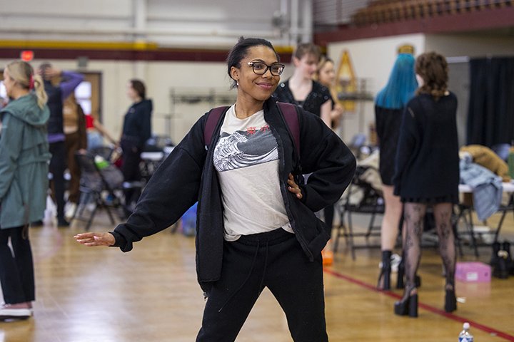 A student-model wearing glasses, a white t-shirt and black sweatshirt practices fashion show poses.
