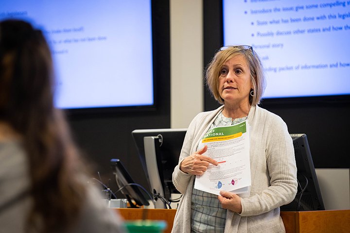 A woman in a light brown sweater stands in front of a group of students in an auditorium while holding a piece of paper and giving instructions.