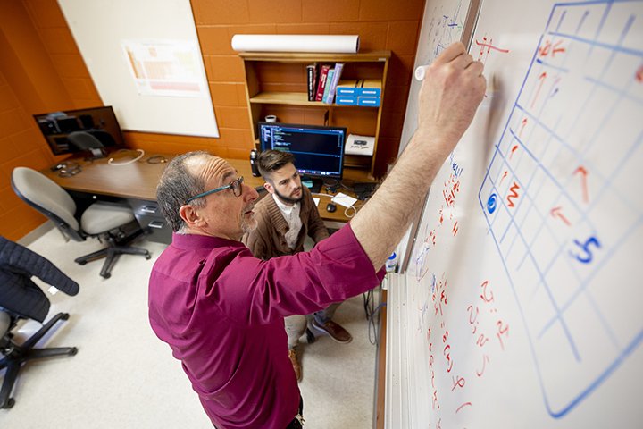 A male professor wearing a wine-colored shirt and glasses, stands at a white board as a student in a brown sweater and beard sits and watches.