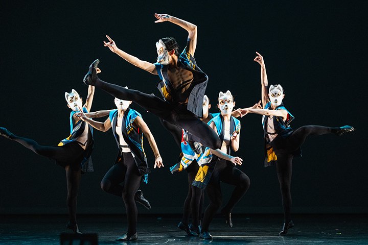 A dance group wearing vests and cat masks performs on a stage with the center dancer leaping in the air.