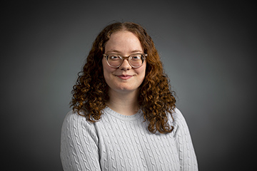 A female student with long hair and glasses wearing a gray sweater smiles at the camera.