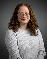 A female student with long hair and glasses wearing a gray sweater smiles at the camera.