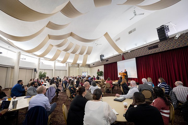 A large group of people sit at tables looking at a presenter behind a podium inside the University Center.