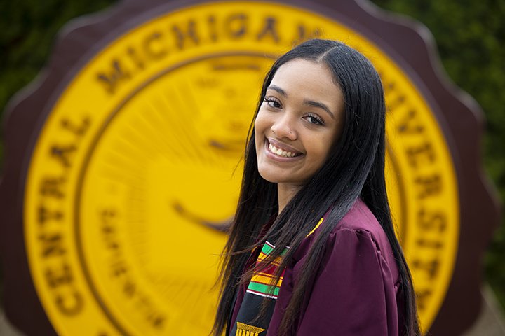 A female student in a graduation gown stands in front of the CMU seal, smiling at the camera.