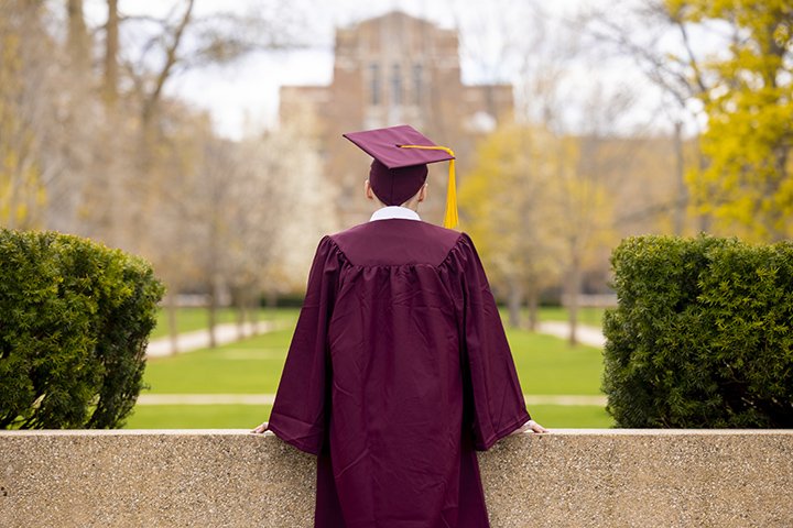 A male college student wearing a graduation cap and gown has his back to the camera while looking at Warriner Hall in the distance.
