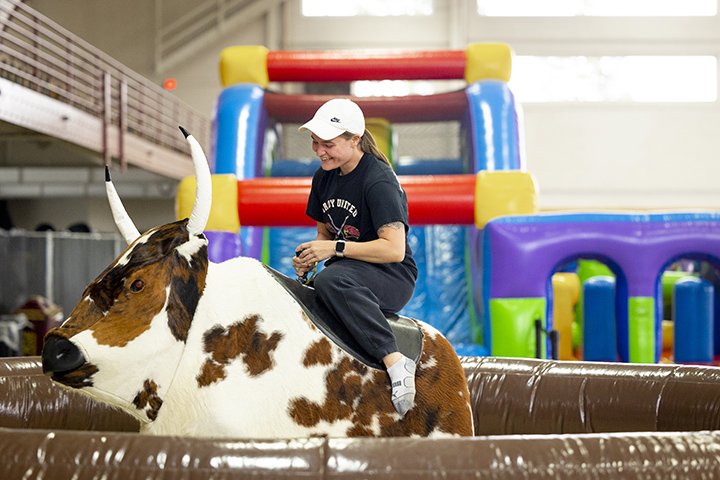 A college student wearing a baseball cap rides a mechanical bull.