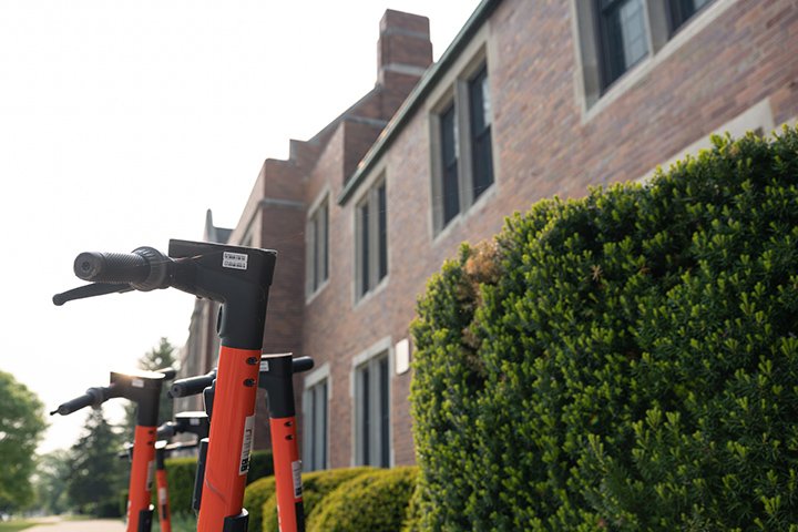 Orange electric scooters sit parked next to shrubs and a brick building.