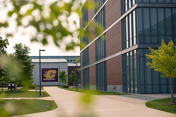 The view looking through tree branches down a sidewalk towards large buildings, once of which features a large CMU logo on its side wall.