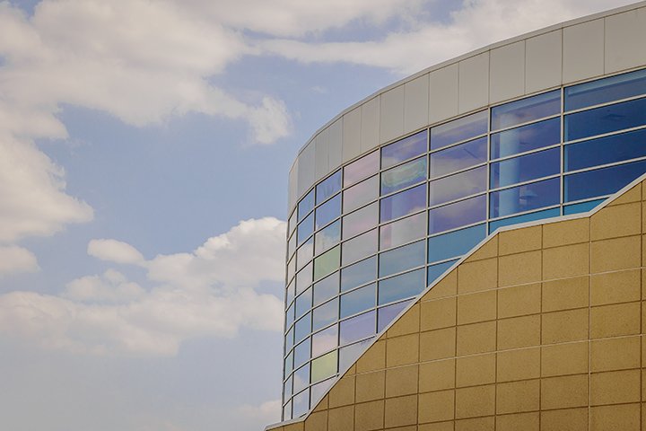 A blue sky with white clouds reflects on the windows of Charles V. Park Library, a light brown building with reflective blue windows.