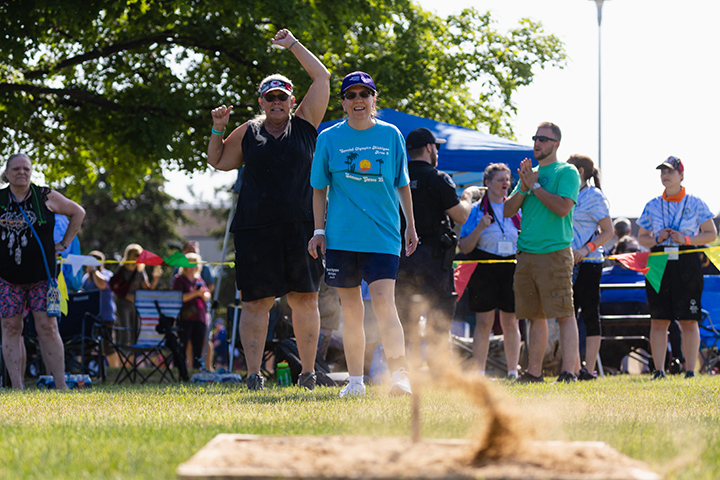 A Special Olympics Michigan athlete in a light blue shirt and a blue hat throws a horseshoe towards a horseshoe pit as spectators cheer.