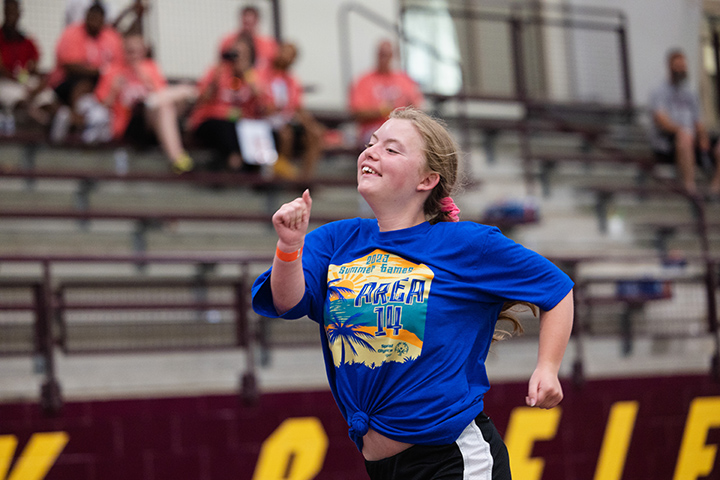 A Special Olympics Michigan athlete in a blue shirt with blonde hair smiles while running down the track inside the IAC.