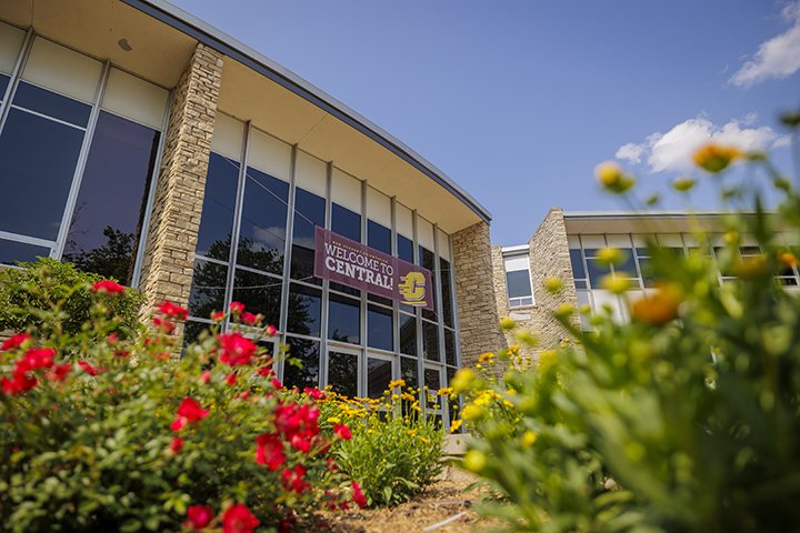Red and white flowers bloom in the foreground of a low angle wide shot of the Bovee University Center.