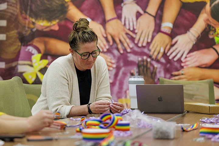 A woman with her hair in a bun and wearing a white sweater sits at a table making pride ribbons.