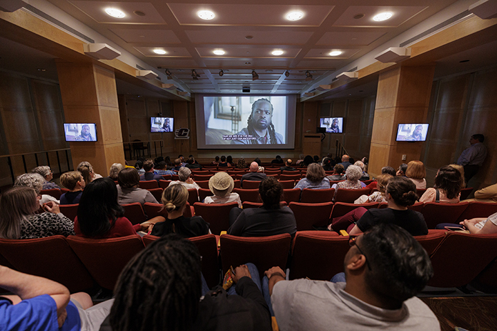 A large group of people sit in an auditorium looking at a movie screen showing a documentary as four televisions flank the sides of the room showing the same documentary.