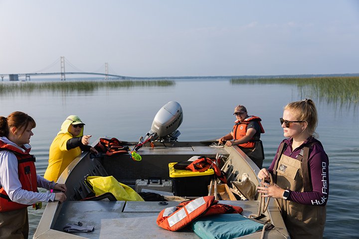 Four people hold onto the sides of a small boat while wearing waders in the water as the Mackinac Bridge stands tall in the distance.