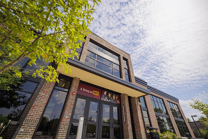 A bright sun shines down on a large brick building with large windows and doors. A green tree stands tall near the entrance.