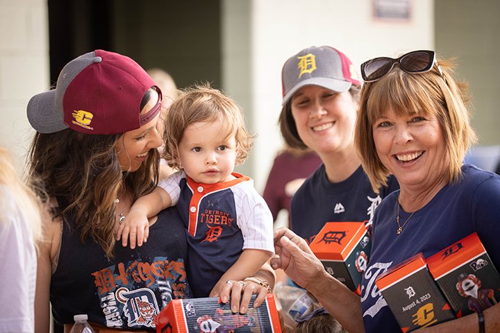 Three women and a small child dressed in Detroit Tigers gear and wearing CMU baseball hats smile at the camera.