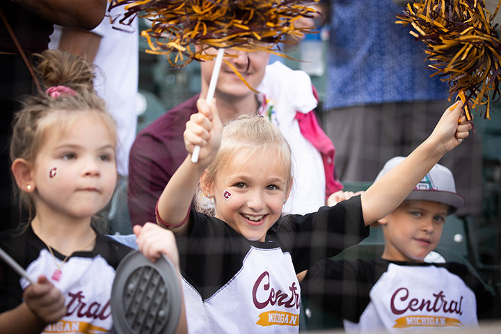 Three young children wearing CMU shirts and temporary face tattoos smile and cheer from the stands at Comerica Park.