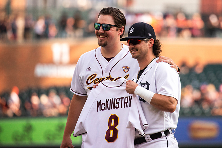 Jake Sabol and Zach McKinstry stand together on a baseball pitchers mound smiling at the camera while McKinstry holds up a white CMU baseball jersey with his last name and 
