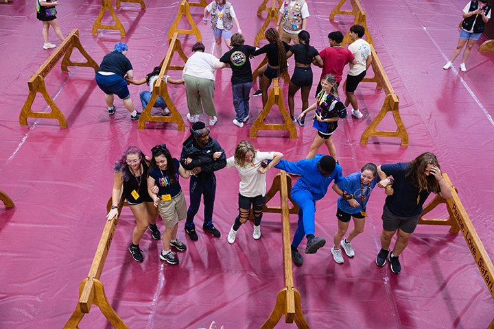 Two groups of students stand arm-in-arm walking together across a maroon-colored floor.