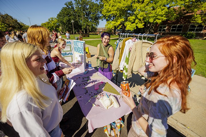 Five female college students stand outside near a table talking to each other during the MainStage event.