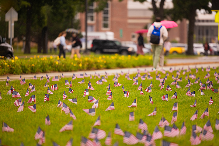 Hundreds of small US flags are planted in the grass near the Park Library as part of a 9/11 tribute. Students walk by on a path in the background.
