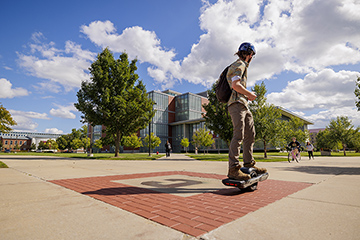 White fluffy clouds float past a blue sky over CMU's campus as a student wearing a backwards baseball hat rides a Onewheel skateboard through the middle of campus.