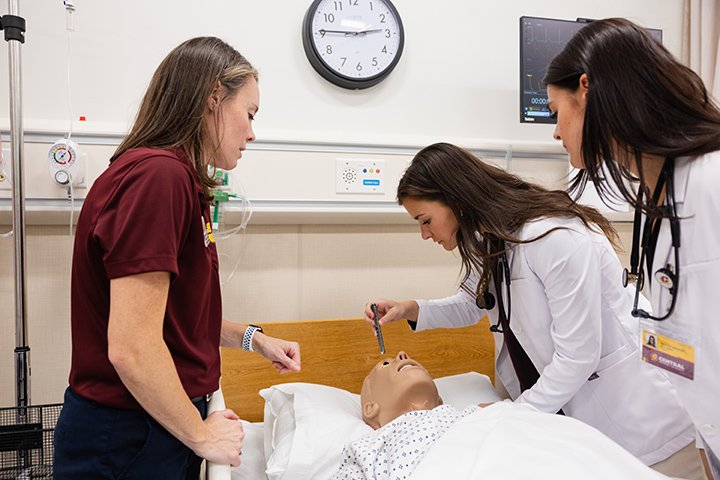 Three female health professions students practice medical procedures on a mannequin inside a hospital room.