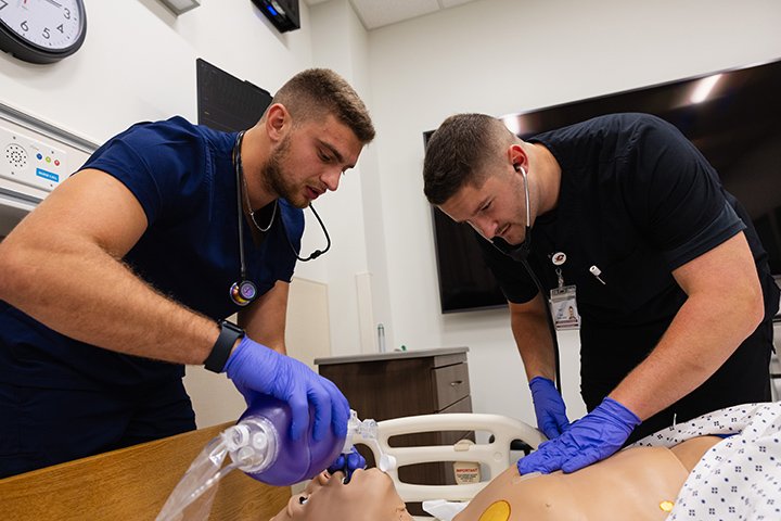 Two male health professions students practice medical procedures on a mannequin inside a hospital room.