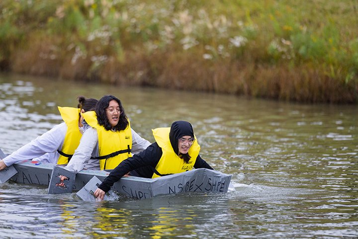 Three students in yellow life vests paddle a cardboard boat in a pond.