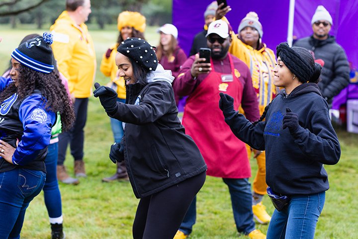 Three young adults in coats and hats dance at the Black Alumni tailgate.