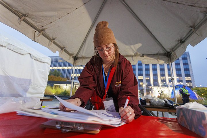 CMU alumni Madi Morell writes on a clipboard while standing at a table underneath a tent.