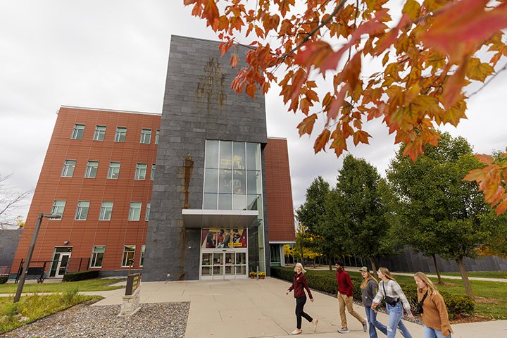 A tour group of five people walk past the Education and Human Services Building as a leafy tree with red and orange leaves stands in the foreground.