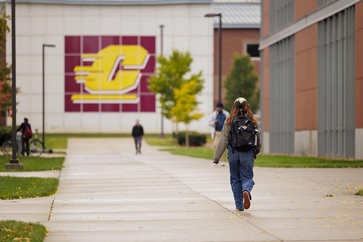 A student wearing a backpack walks down a sidewalk away from the camera as a giant CMU action C logo on the side of the Engineering and Technology Building is in the distance.
