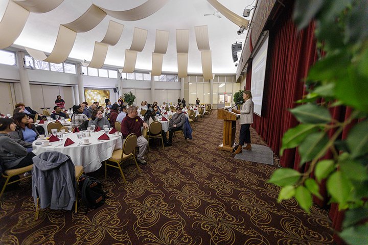In the brightly lit Rotunda Room, dozens of people sit at round tables enjoying brunch while a presenter stands at a podium speaking.