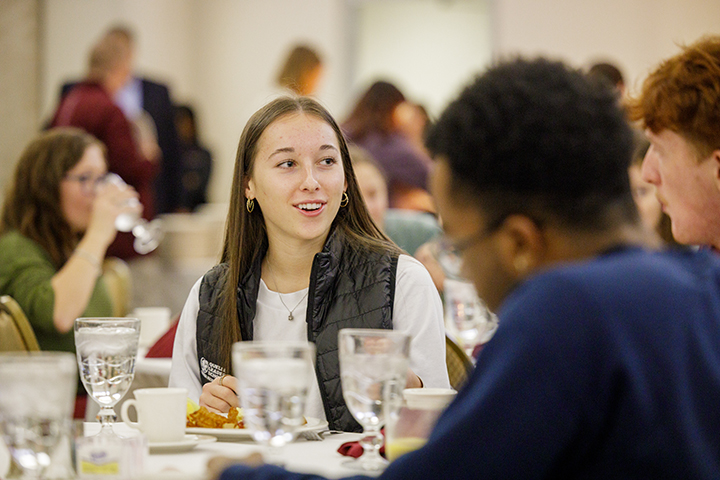 A female college student sits at a table, smiling while in conversation with others at her table while they all enjoy brunch.