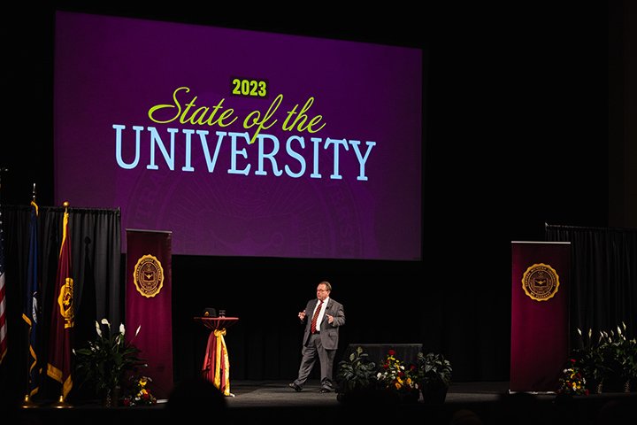 CMU President Bob Davies stands on stage in an auditorium as a large screen in the background reads