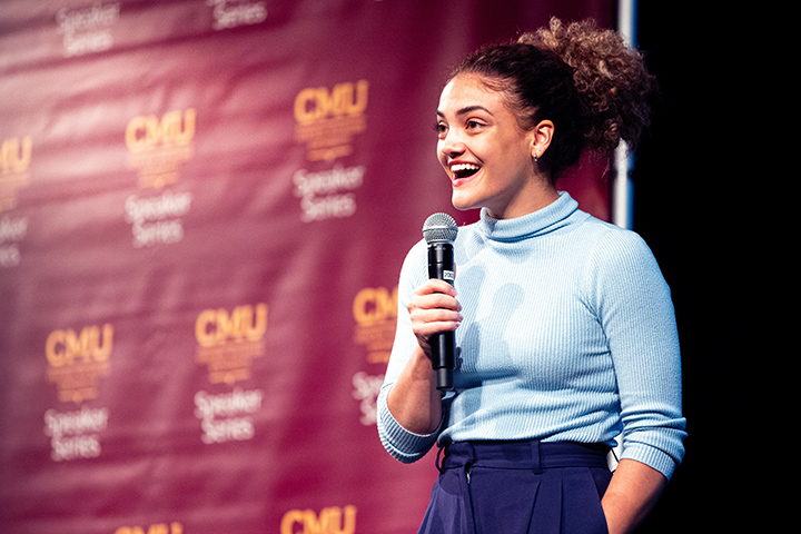 Former Olympic gymnast Laurie Hernandez wears light blue shirt while holding a microphone and speaking to a crowd. A CMU step and repeat banner stands behind her.