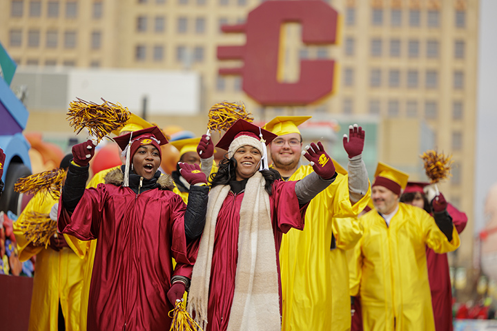 A group of parade marchers dressed in commencement attire walks in downtown Detroit next to a CMU float.