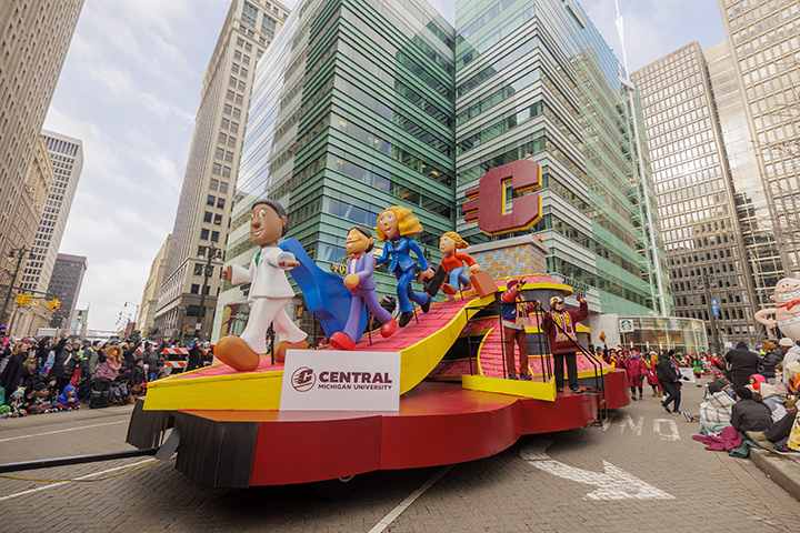 CMU's colorful parade float travels through downtown Detroit between tall buildings.