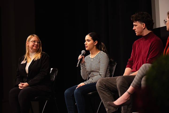 A female student in a striped sweater holds a microphone while sitting on stage surrounded by other students as part of a panel discussion in an auditorium.