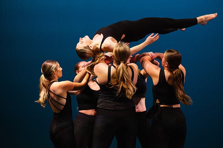 Five female dance students dressed in black hold another dance student in the air.
