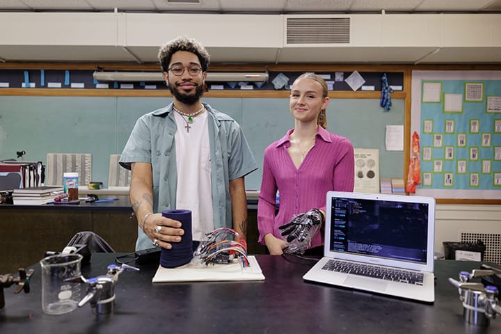 A male student with glasses and a beard and a female student in a dark pink short stand at a table containing a laptop and textile sensors.