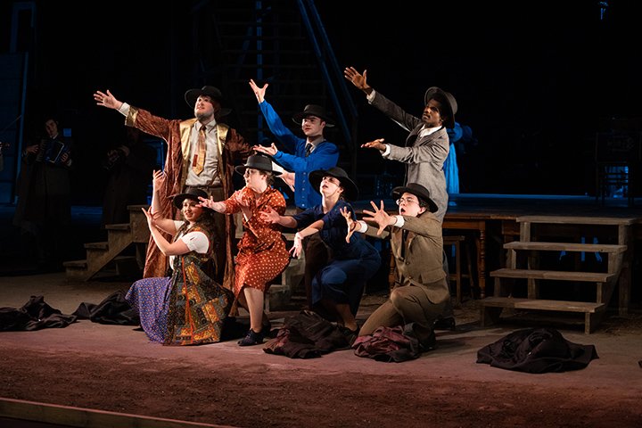 Seven theatre students wearing dresses and suits look off to the distance while reaching their arms out to the spotlight coming from the back of the auditorium.