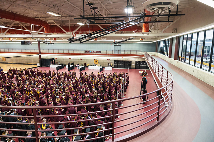 A top-down view from the upper track surrounding basketball courts in the SAC, President Davies addresses soon-to-be graduates dressed in maroon-colored caps and gowns.