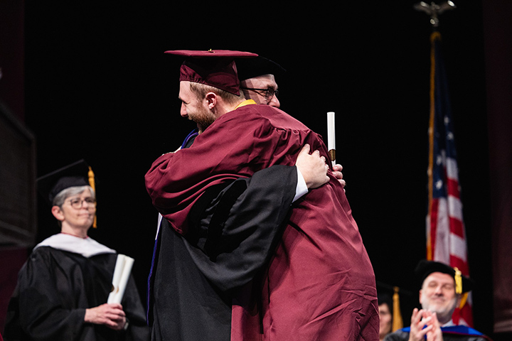 A CMU graduate in a maroon cap and gown hugs President Davies on stage after receiving his diploma.