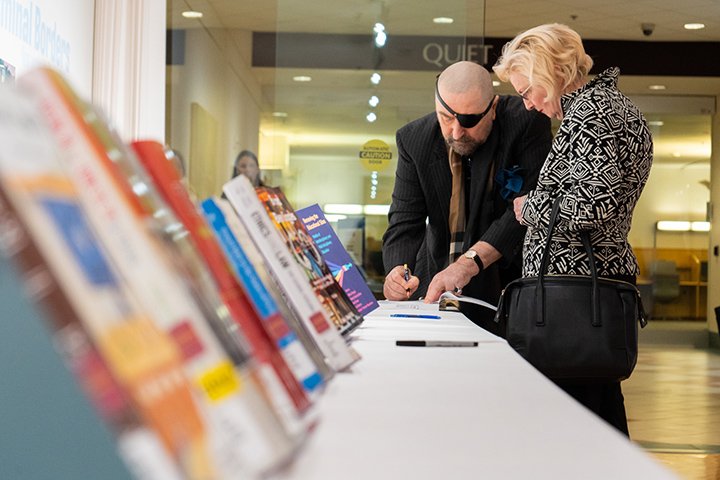 A man signs a book as a woman looks on. Both stand next to a table featuring recently-published books by CMU faculty members.