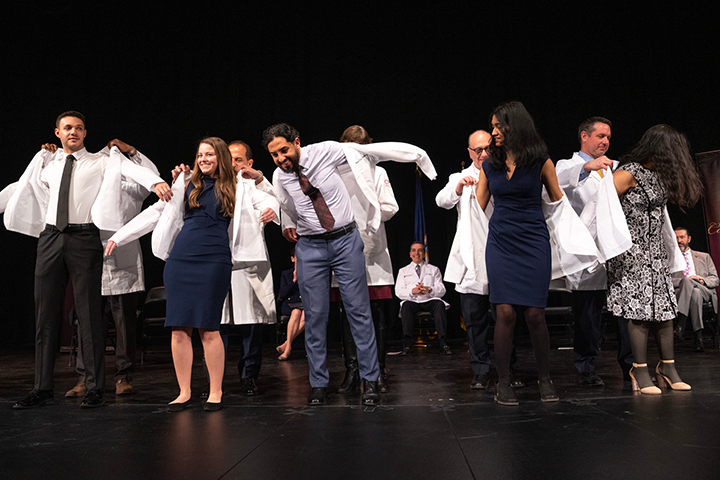Medical students stand on a stage putting white medical coats on.